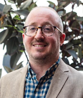Man with glasses wearing a blue and white flannel shirt with a gray jacket.