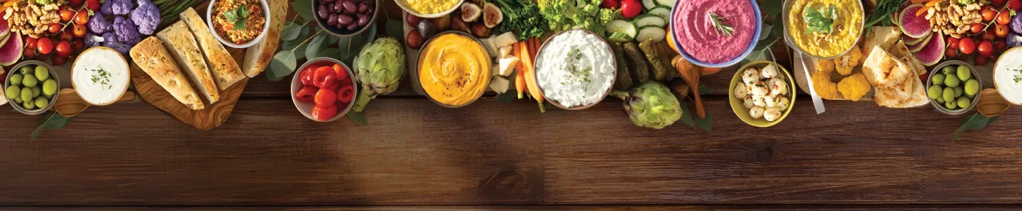 nourish-bg-desktop.webp Large array of vegetables and spreads on wooden surface.
